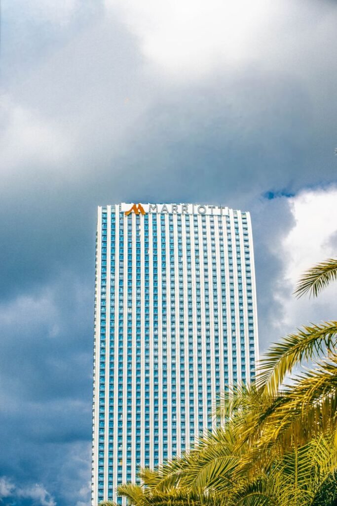 A modern Marriott skyscraper stands tall against a dramatic, cloudy sky, framed by palm fronds.